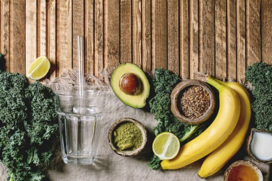 Glass for green healthy vegan smoothie. glass straw. Ingredients above. Kale. bananas. avocado. lime. non-diary milk. honey. matcha powder and seeds over wooden plank cloth background. Flat lay. space
