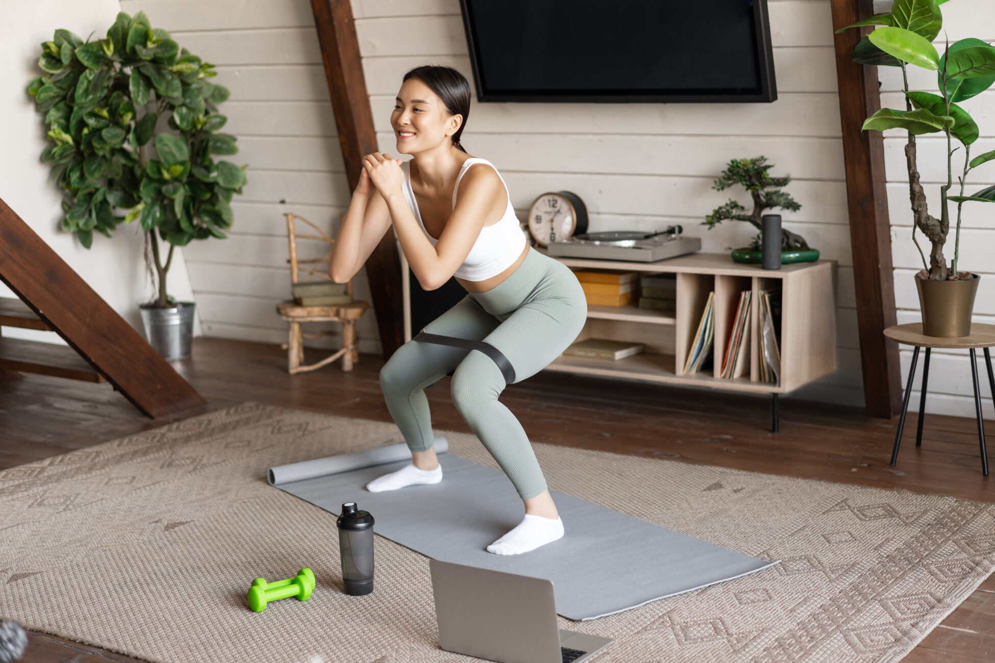 Cute asian fitness girl at home doing workout, squats with stretching elastic rope on legs, standing on floor mat, listening to online sport instructor on laptop video