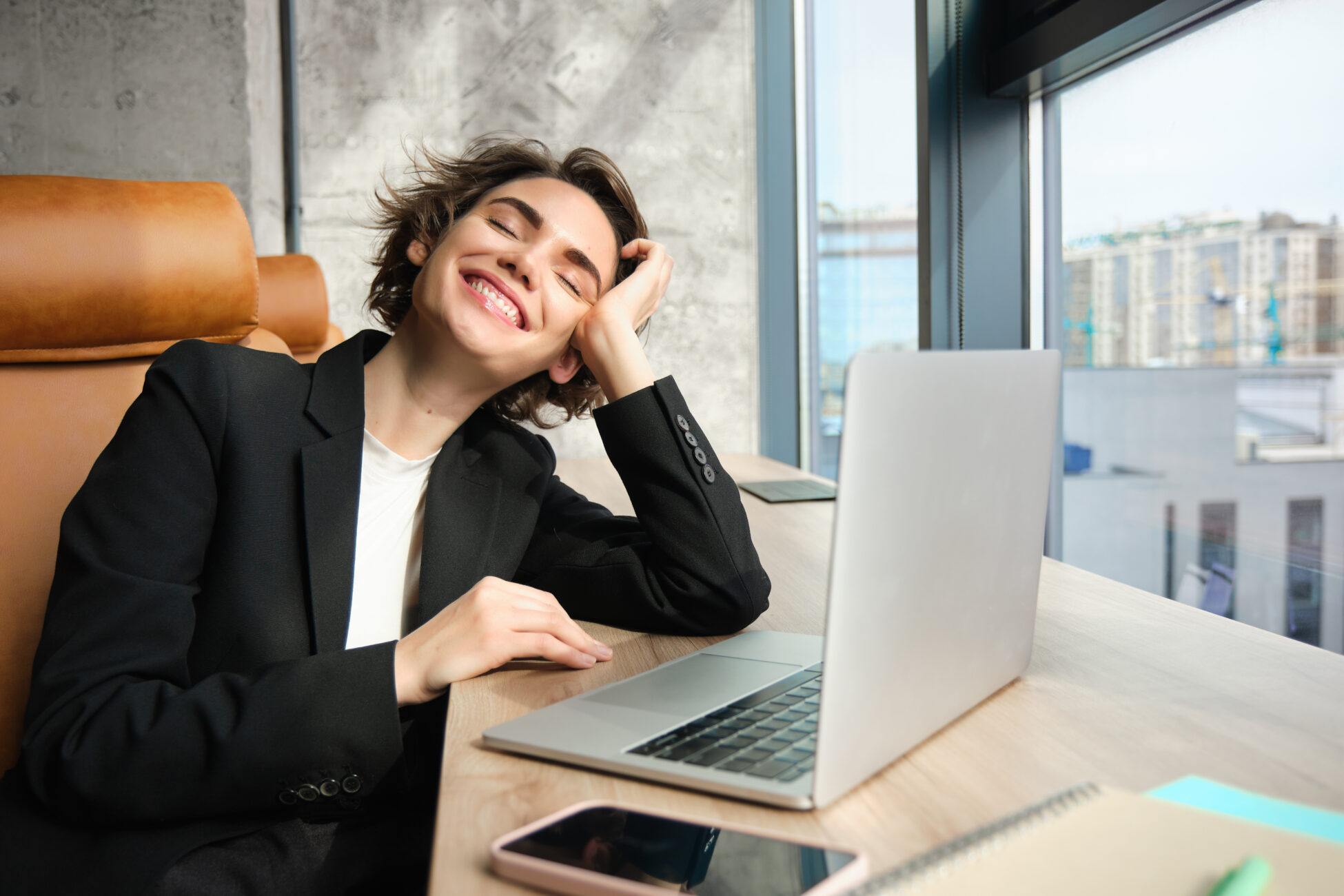 Portrait of happy young female manager, company ceo working in office. Businesswoman with carefree smile, sitting in front of laptop, looking outside window