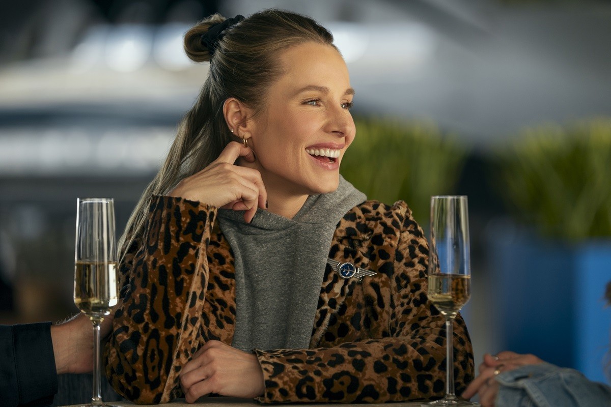 "Woman in leopard print jacket sitting at a table, smiling and talking with others, two glasses of champagne in front of her, modern indoor setting with blurred background and plants."