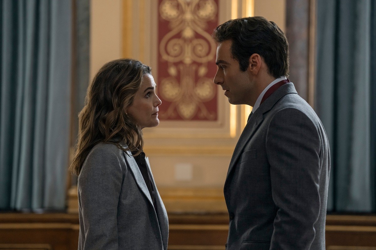 "Two people in business attire stand facing each other in an ornate, formal room with blue curtains and decorative wall panels, appearing engaged in a serious conversation."